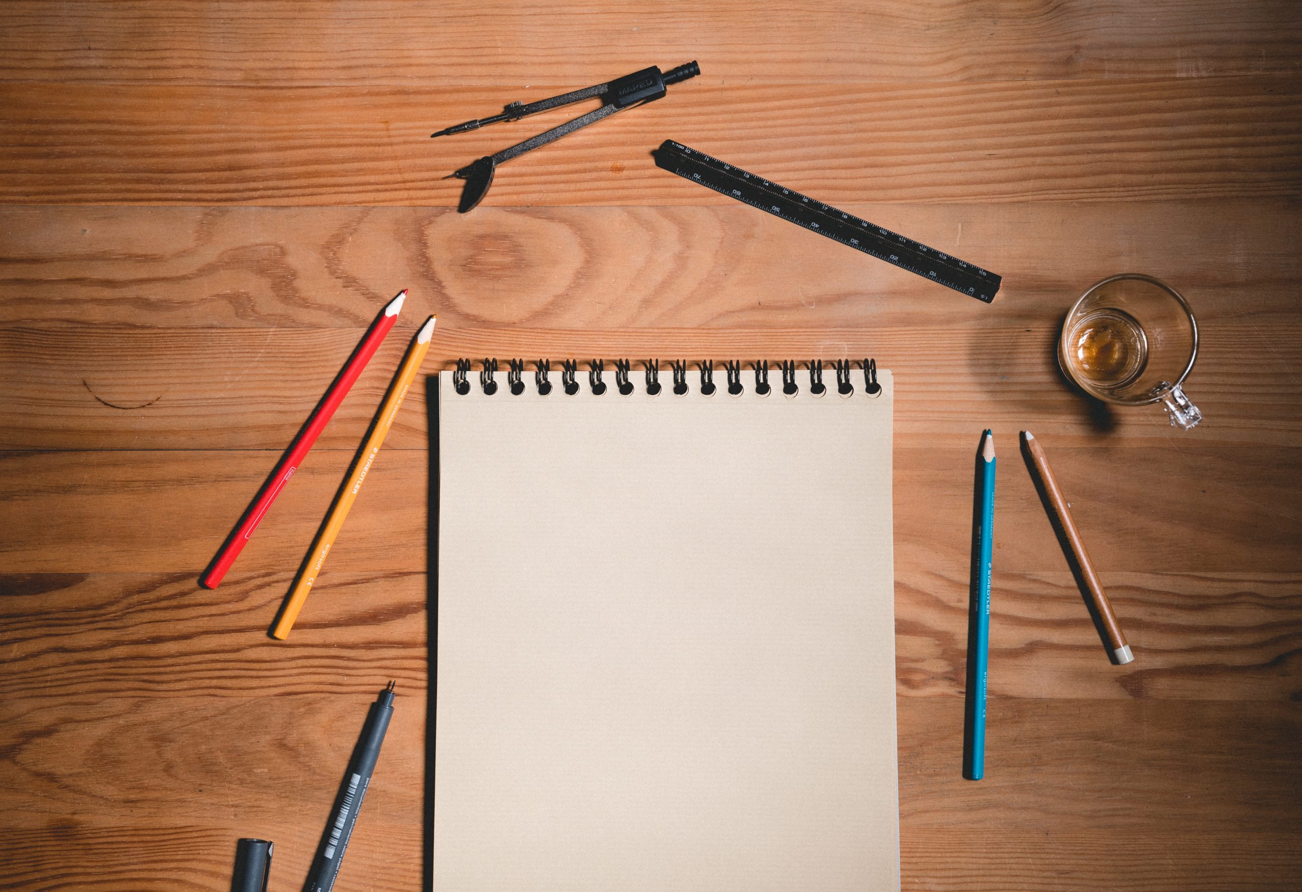 A picture of a notebook lying on a wooden desk. Around the notebook is color pencils, pens, ruler, a compass ruler and an empty cup.